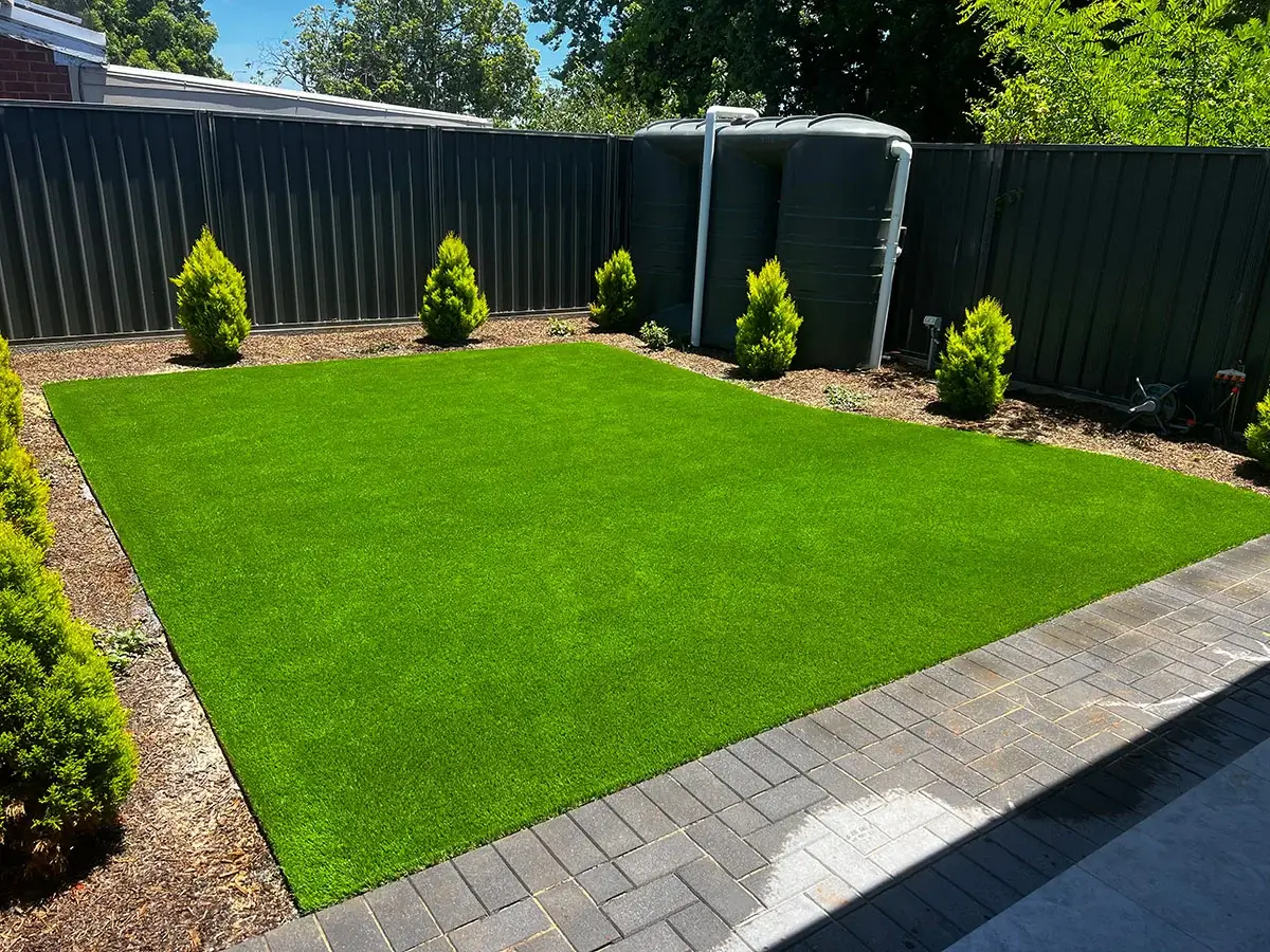 Backyard with vibrant artificial turf, bordered by small evergreen shrubs and black metal fencing, with water tanks visible in the corner.