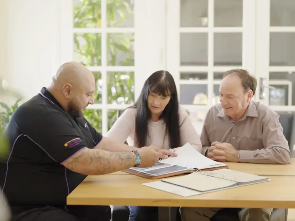 Consultant discussing documents with a middle-aged couple at a table in a bright room with large windows in the background.