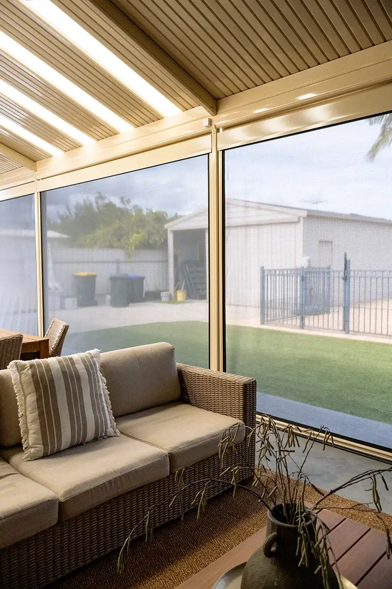 Comfortable outdoor lounge area with Patio Blinds, featuring a wicker sofa with striped cushions, and a view of the backyard with a storage shed and grass.