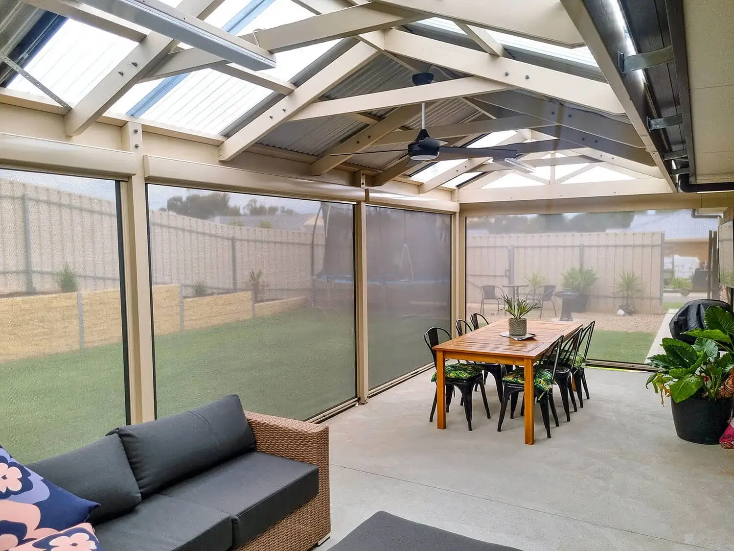 Spacious outdoor area with Outdoor Patio Blinds, featuring a dining table with black chairs, a wicker sofa with floral cushions, and potted plants, all under a modern pergola.