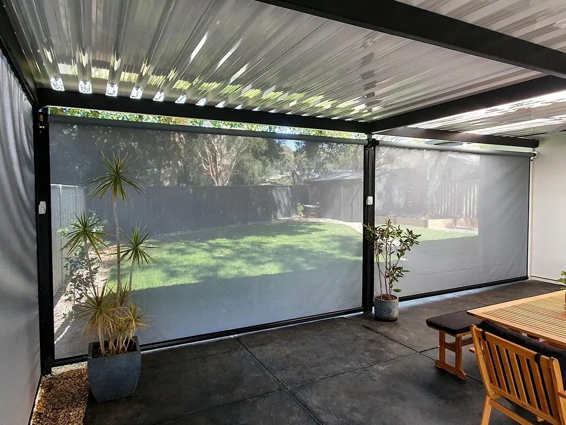 Covered patio area with outdoor blinds, providing shade and privacy, featuring potted plants and a wooden dining set, with a view of the backyard.
